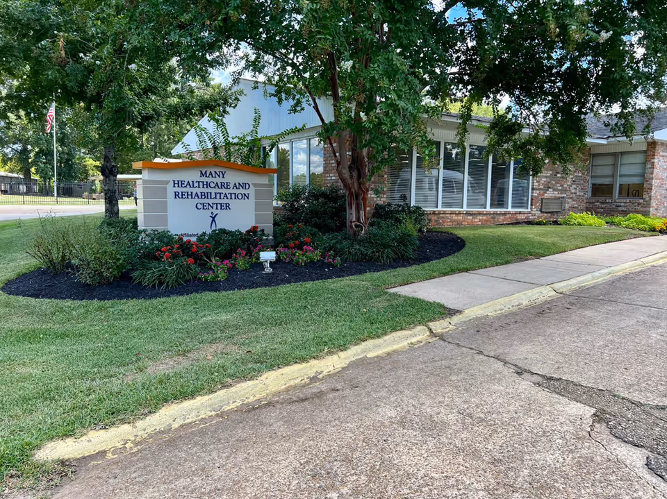 Sign for Many Healthcare and Rehabilitation Center on a landscaped lawn in front of a brick building with trees and windows.