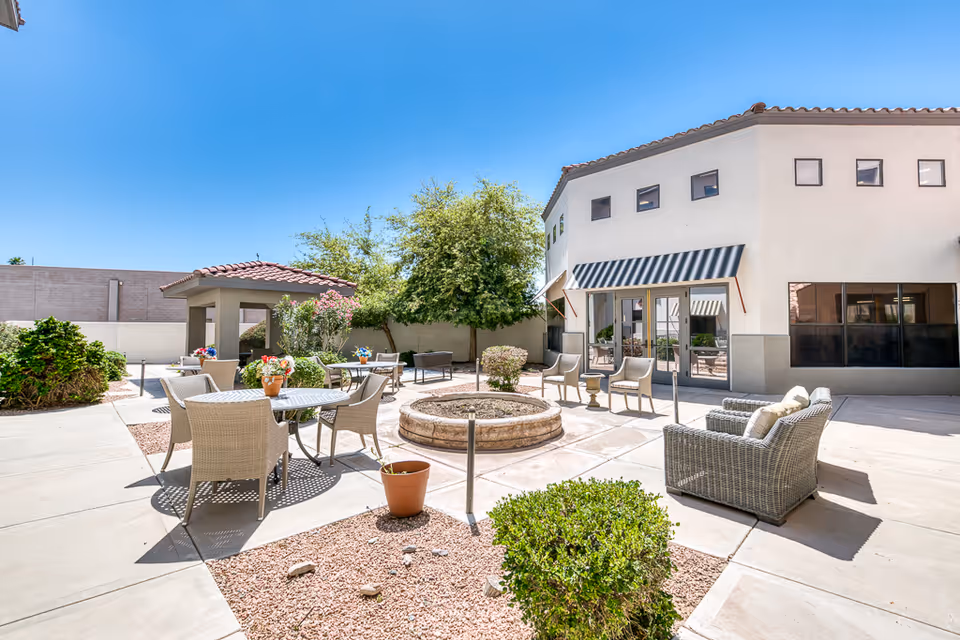 Outdoor patio area at The Woodmark at Sun City with multiple seating arrangements including wicker chairs and tables, a central circular planter, potted plants, and a small covered gazebo. The building has a white exterior with a striped awning over the glass doors, and the sky is clear and blue.