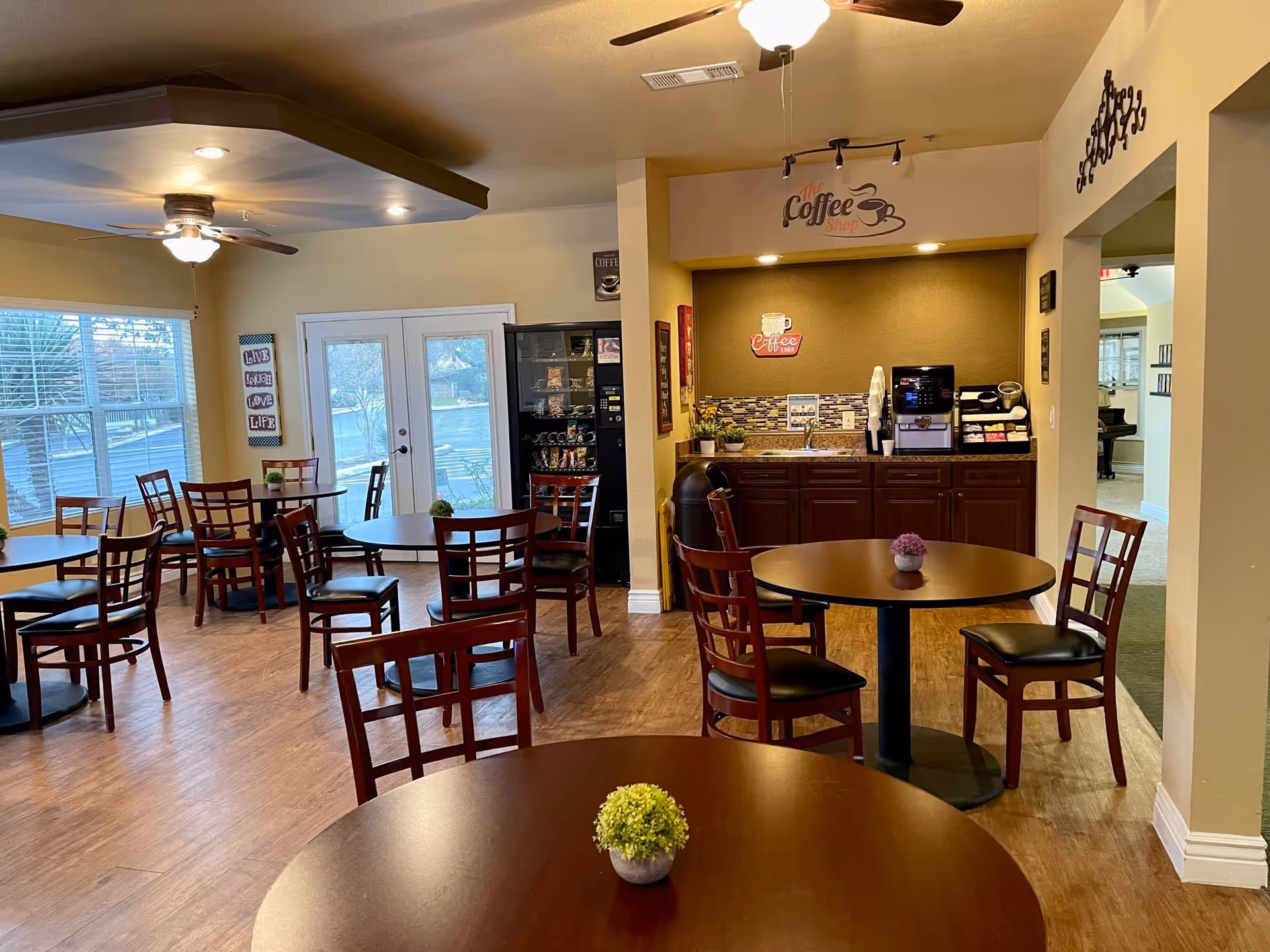 Interior view of a cozy coffee shop area in a retirement community with several round wooden tables and chairs. There is a coffee station with a coffee machine, cups, and condiments against the wall. Large windows allow natural light to brighten the room, and decorative signs related to coffee and positive messages are displayed on the walls.