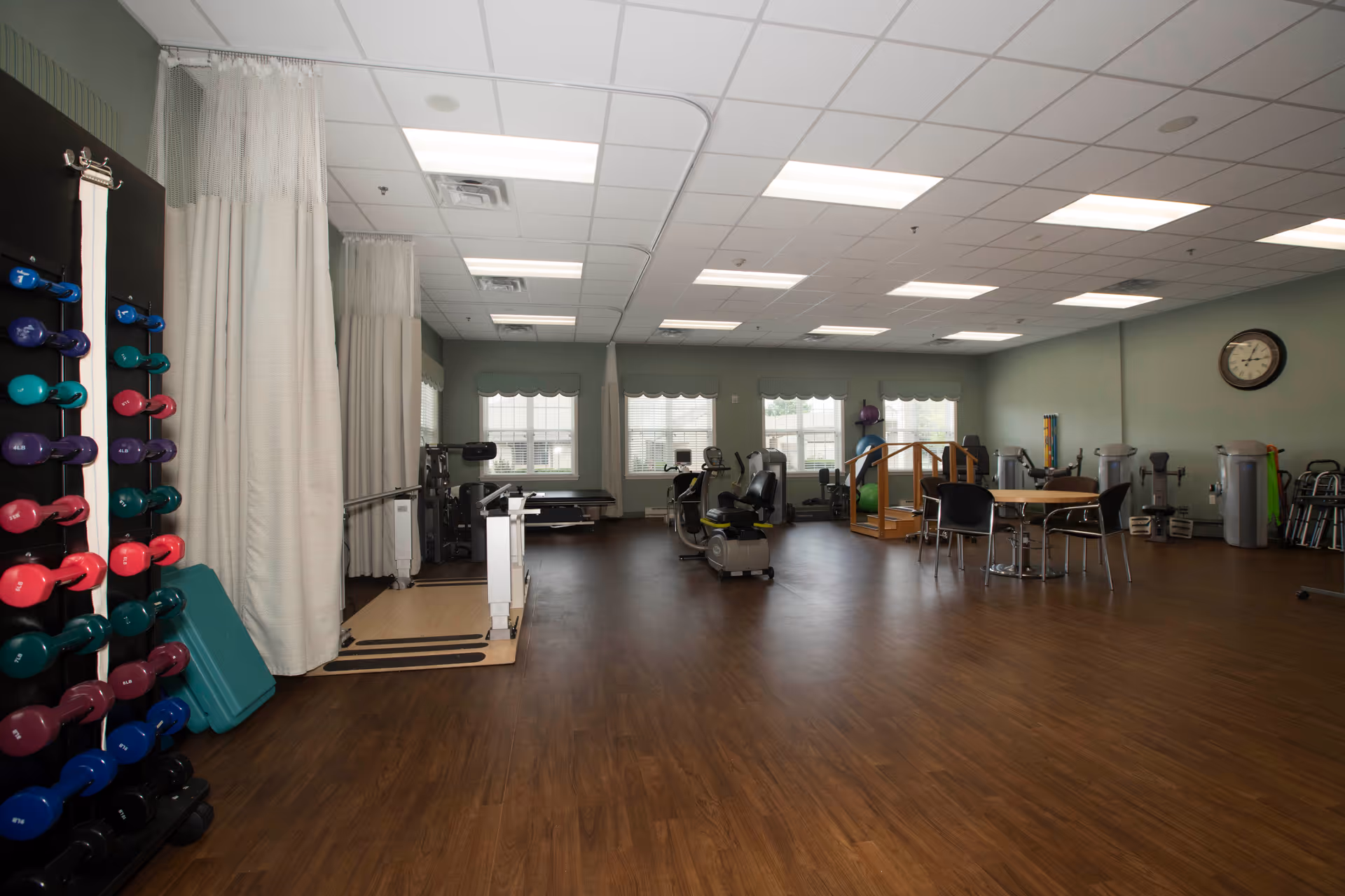 A spacious exercise room with wooden flooring and light green walls. The room contains various exercise equipment including colorful dumbbells on a rack, stationary bikes, weight machines, and exercise balls. There are several windows with white blinds letting in natural light, a round table with chairs, and a clock on the wall.