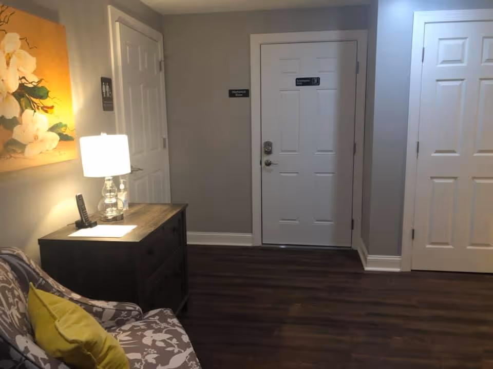 Small seating area with a patterned armchair, side table with a lamp and phone, floral wall art, and three white doors down a hallway.