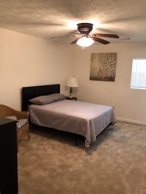 A simple bedroom with a bed covered in a gray bedspread, two pillows, a wicker chair with a cushion, a small nightstand with a lamp, a ceiling fan with a light, a window with blinds, and a framed artwork on the wall.