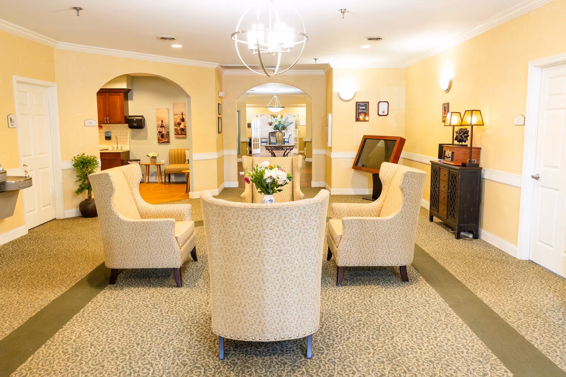 A cozy senior living common area with four beige upholstered armchairs arranged around a small table with a flower vase. The room has light yellow walls, patterned carpet, and soft lighting. In the background, there is an archway leading to another room with additional seating and a kitchenette area. Decorative elements include framed pictures on the walls and a small cabinet with a lamp and vintage radio.