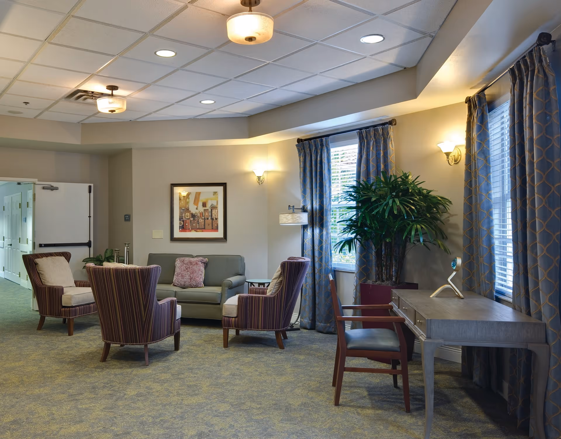 A cozy common lounge with armchairs, a sofa, a desk and a large potted plant beside curtained windows.