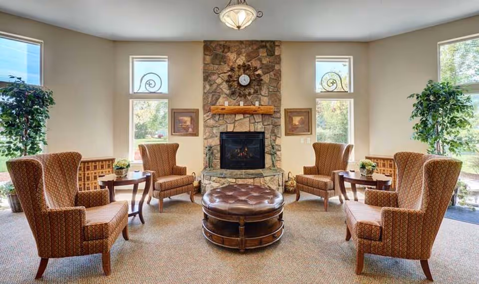 Cozy assisted-living lounge with a stone fireplace, four wingback chairs arranged around a central leather ottoman, side tables and large windows.