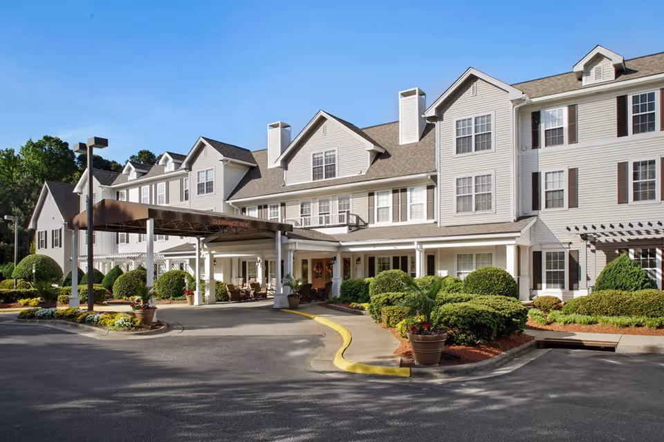 Exterior view of a large, multi-story senior living facility with white siding and multiple windows. The entrance features a covered drop-off area with a brown canopy and well-maintained landscaping including trimmed bushes and potted plants. The sky is clear and blue.