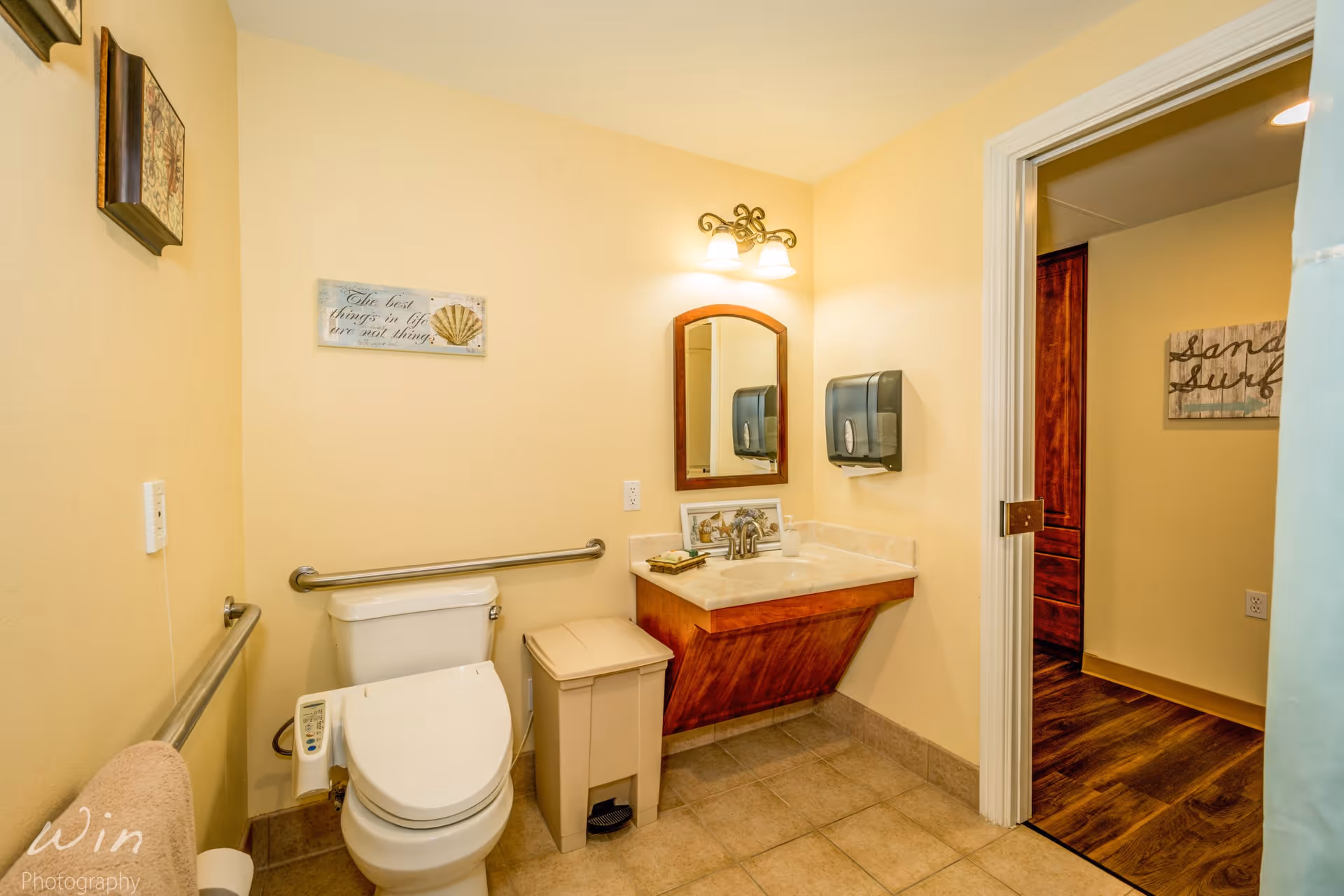 A clean and accessible bathroom with beige walls and tiled floor. The bathroom features a white toilet with a safety grab bar on the wall behind it and another on the left side. There is a beige trash bin next to the toilet. A wooden vanity with a marble countertop and a sink is mounted on the right wall, with a mirror above it and a two-light fixture. A wall-mounted paper towel dispenser is next to the mirror. The bathroom door is open, showing a hallway with wooden flooring and a wall decoration that reads 'sand surf'.