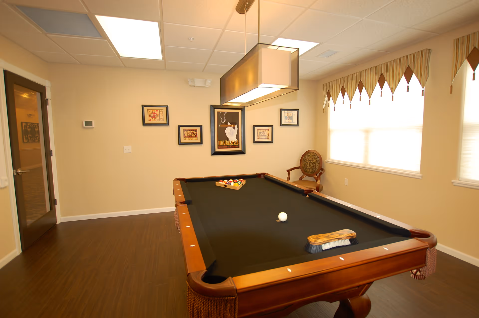 A well-lit room with a black felt pool table in the center, a wooden chair with an ornate backrest near the window, framed artwork on the beige walls, and a rectangular ceiling light fixture above the pool table. The room has wood flooring and windows with striped valances.