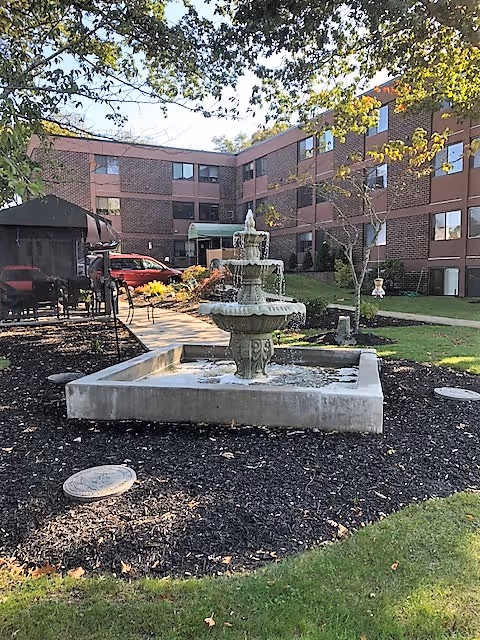 Outdoor courtyard area of a senior living facility with a three-tiered water fountain in the center, surrounded by mulch and grass. In the background, there is a three-story brick building with multiple windows and a covered entrance. Trees with green leaves partially frame the scene.
