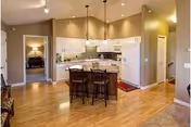 Spacious kitchen area with white cabinets, a central island with three bar stools, pendant lighting, and hardwood floors. Adjacent rooms and hallway are visible, with warm beige walls and recessed ceiling lights.