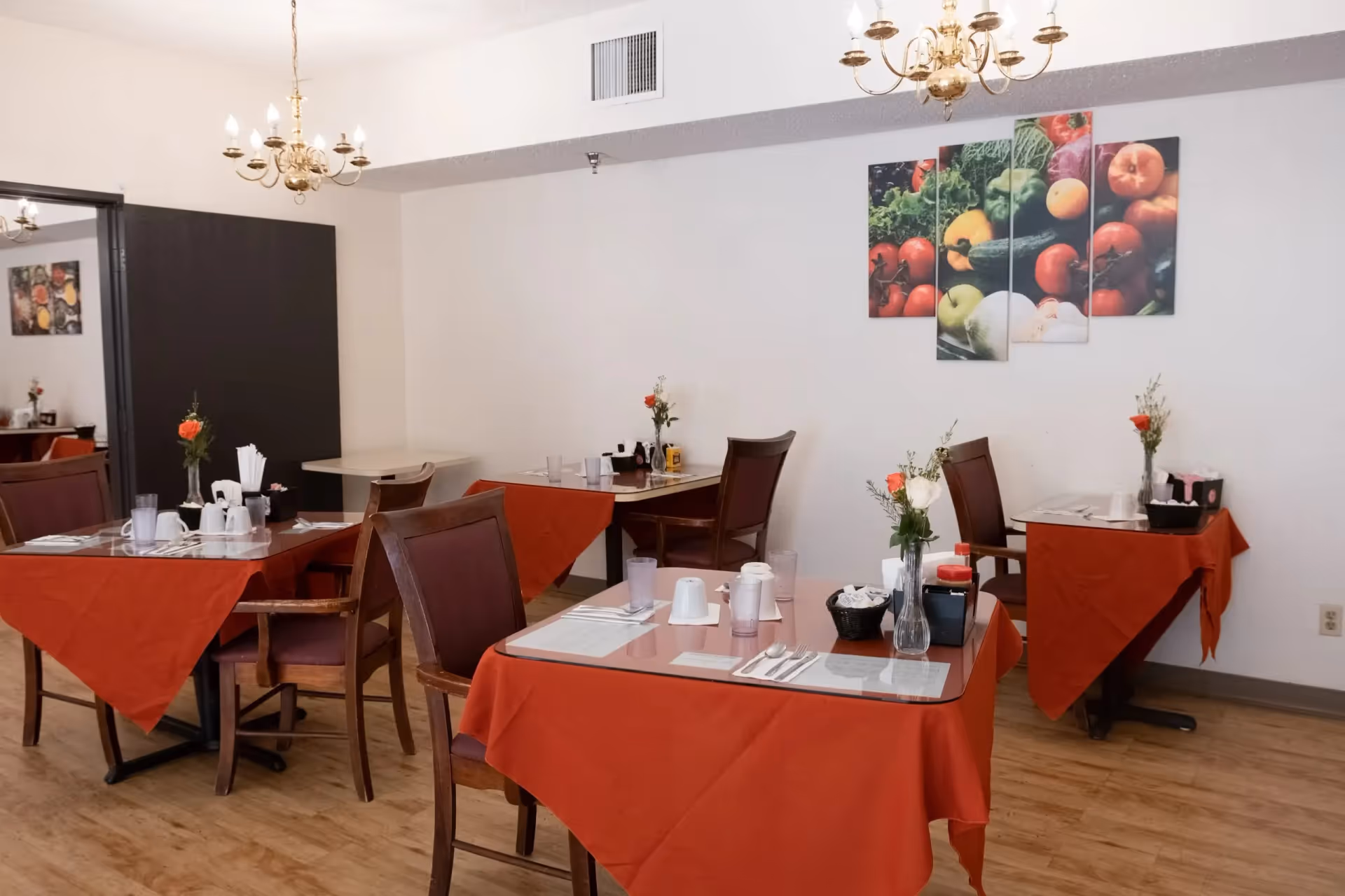 Dining room with several tables covered in red tablecloths and set with placemats, utensils, cups, and small flower vases. The room has wooden chairs, two chandeliers hanging from the ceiling, and a multi-panel wall art featuring various fresh vegetables.