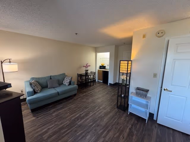 Interior view of a living room area with a light blue sofa with pillows, a floor lamp, a small table with two chairs, and a tall lamp with a square shade. There is a small white shelf with a basket and a door on the right side. The floor is dark wood, and there is an open doorway leading to a kitchen area.