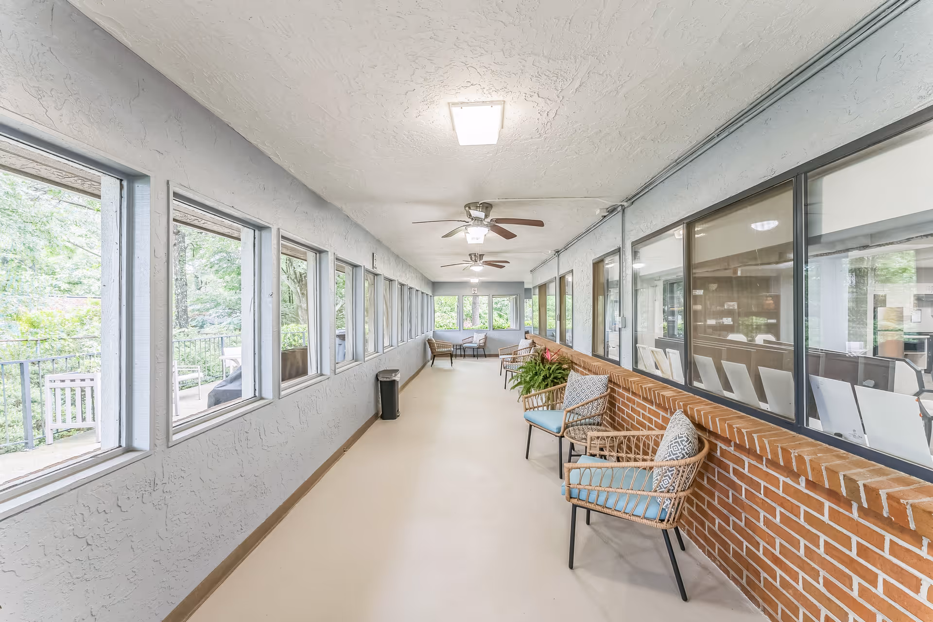 A bright, enclosed corridor with multiple windows on the left side showing an outdoor patio area with benches and greenery. On the right side, there are large interior windows looking into another room with chairs. The corridor has a beige floor, textured white ceiling with ceiling fans, and a brick half-wall with several wicker chairs with blue cushions and patterned pillows along it. There is also a small table with a plant on it.