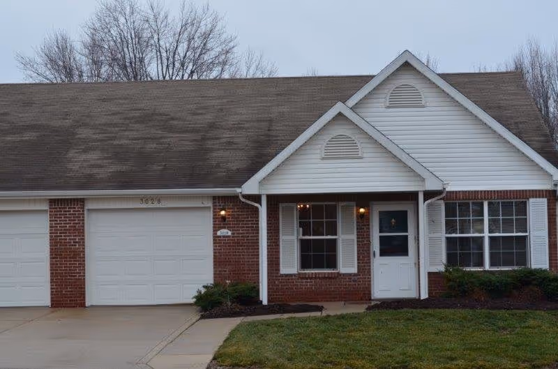 Exterior view of a single-story brick and white siding building with a two-car garage, a front door, and two windows. There is a small lawn and some bushes in front of the building, and leafless trees in the background.