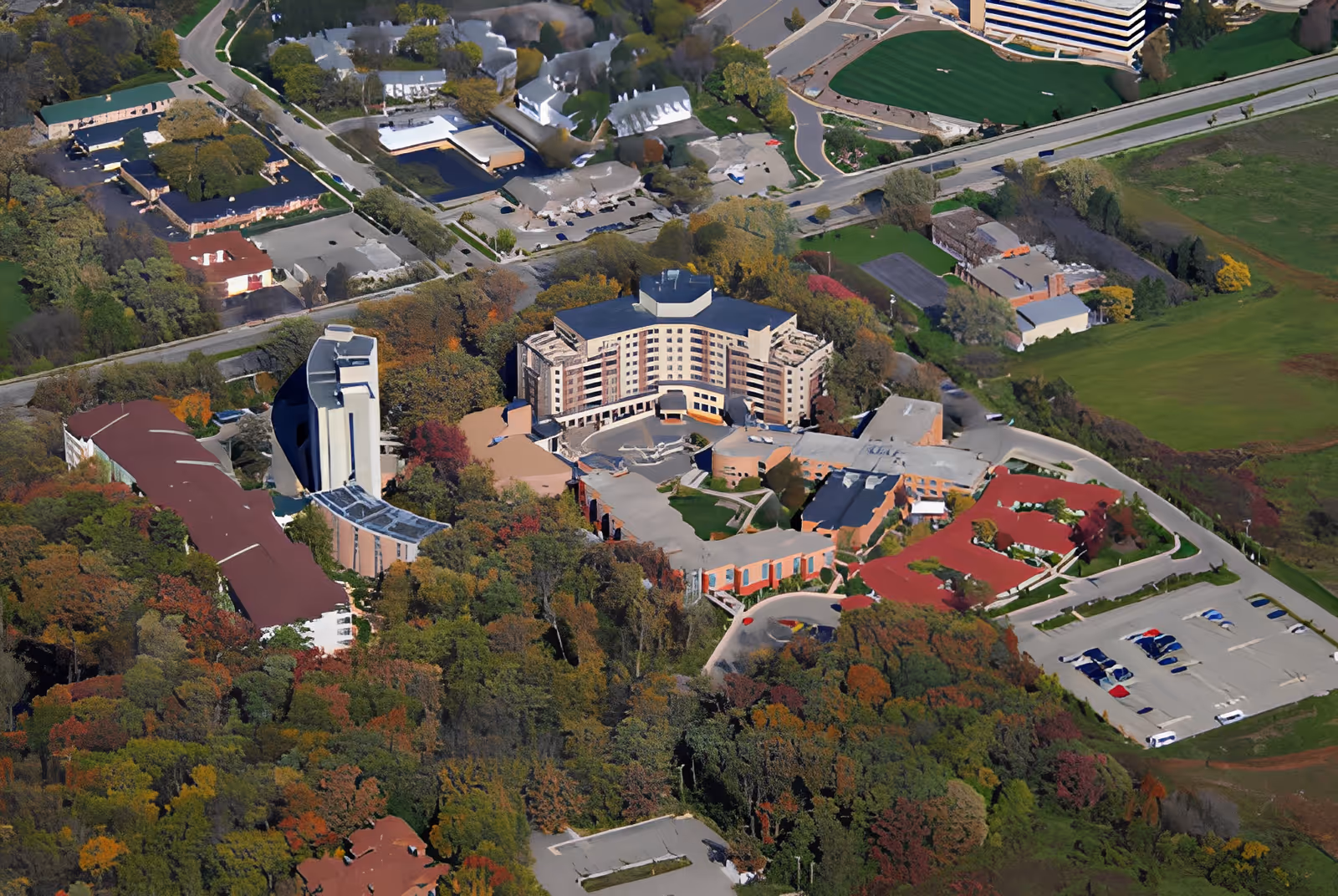 Aerial view of Oakwood Village University Woods facility surrounded by trees with autumn foliage, parking lots, and nearby roads. The facility consists of multiple connected buildings with varied roof styles and colors, set in a green, suburban area.