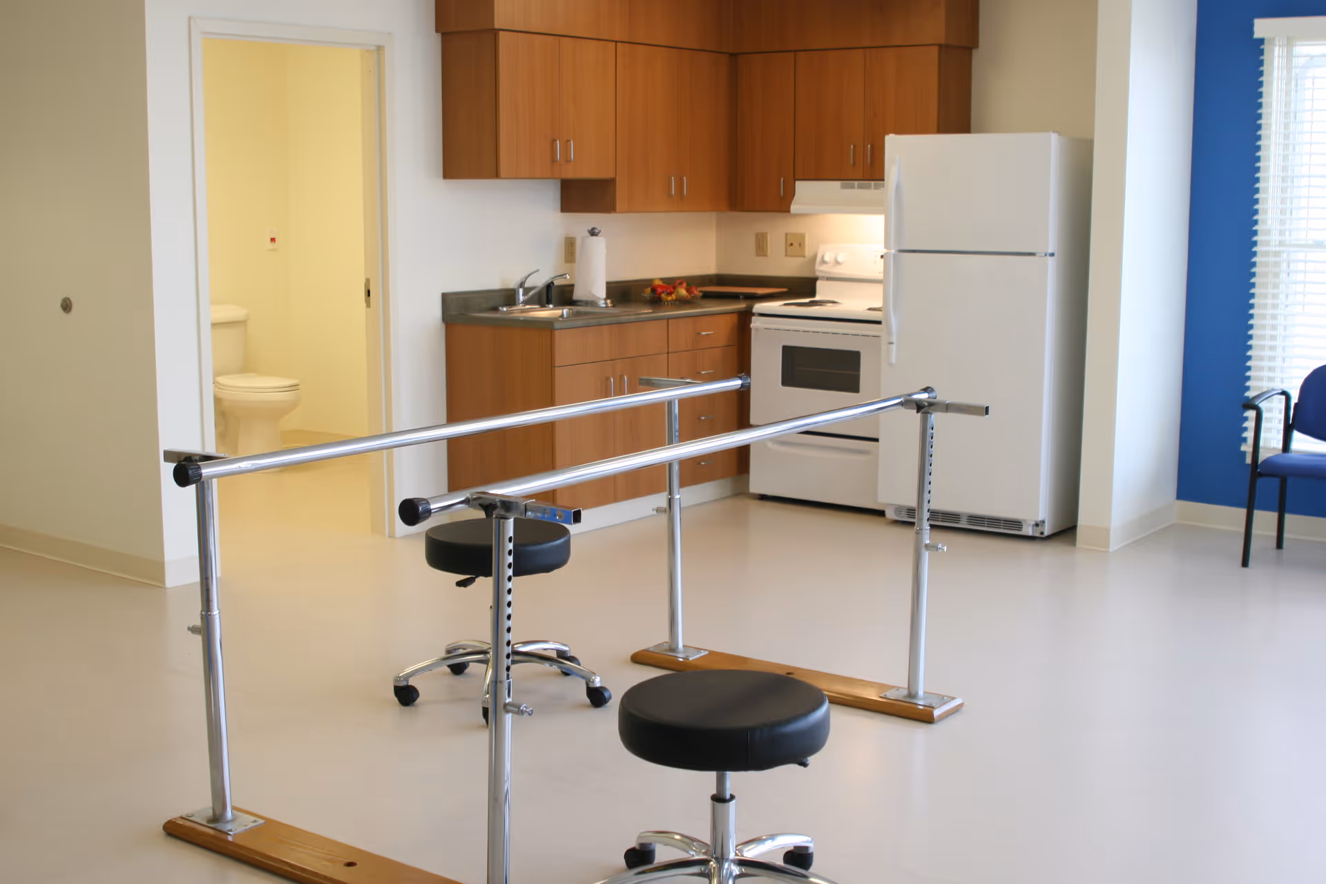 A room with a small kitchen area featuring wooden cabinets, a sink, a white stove, and a refrigerator. In the foreground, there are parallel bars used for physical therapy and two black stools on wheels. To the left, there is an open door revealing a bathroom with a toilet. A blue chair is visible near a window with blinds on the right side of the room.