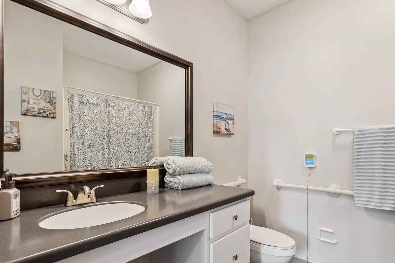 A clean and modern bathroom featuring a large mirror above a dark countertop with a built-in sink. On the countertop, there are two neatly folded towels, a candle, and a soap dispenser. The bathroom has a white toilet with grab bars on the wall beside it, a towel rack with a striped towel, and a shower curtain with a decorative pattern. The walls are painted light beige and there are two small framed pictures hanging on the walls.
