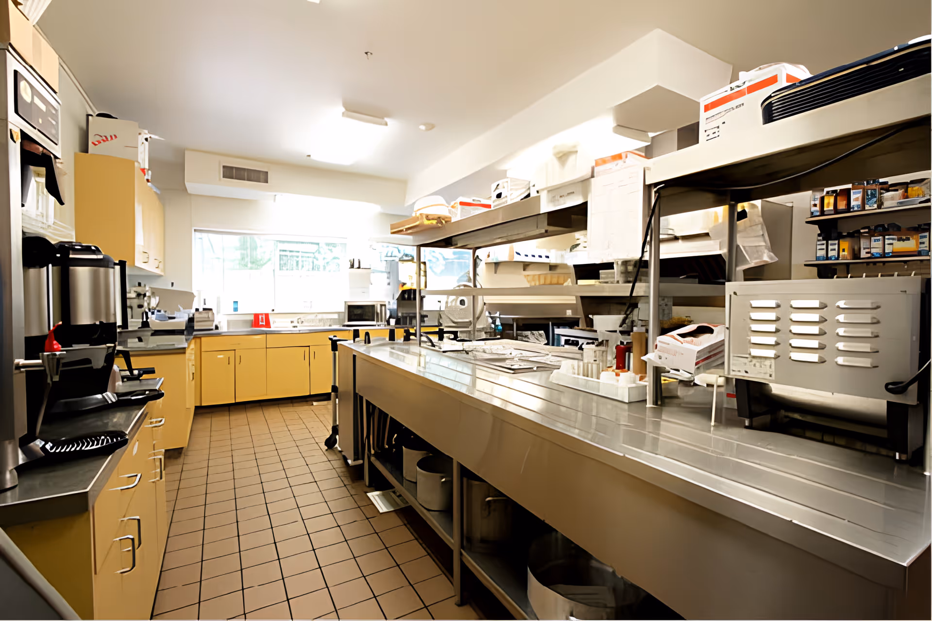 A commercial kitchen with stainless steel countertops and shelves, yellow cabinets, various kitchen appliances, and a tiled floor. The kitchen is well-lit with natural light coming through a window at the far end.