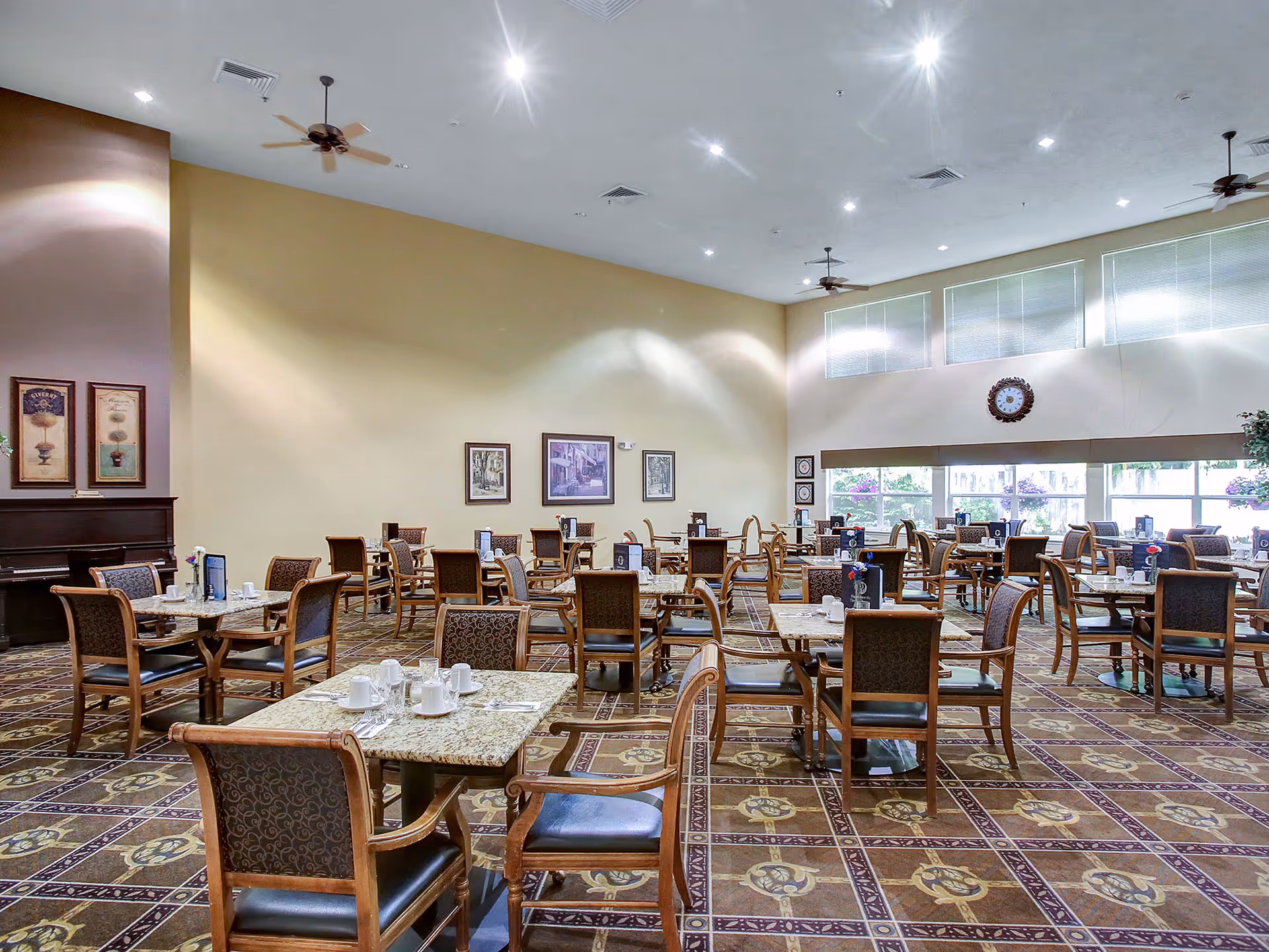 A spacious dining room with multiple tables and chairs arranged neatly. Each table has place settings including cups, saucers, and utensils. The room features large windows allowing natural light to enter, ceiling fans, framed artwork on the walls, and a patterned tile floor.