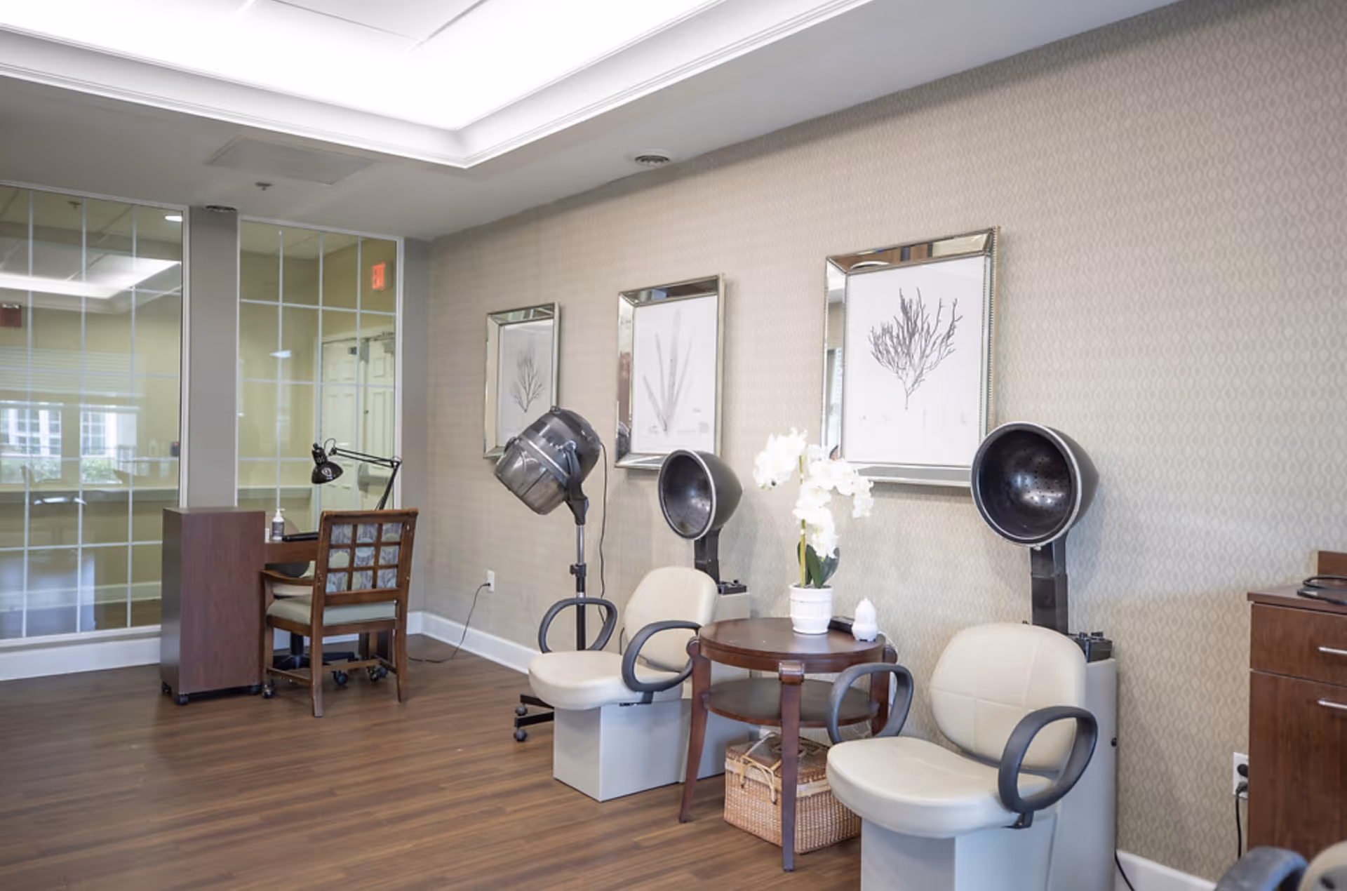 Interior view of a salon area in a senior living facility with two hair drying chairs, a small round table with a white flower pot, framed botanical prints on the wall, and a manicure station with a wooden chair and desk lamp near glass-paneled walls.