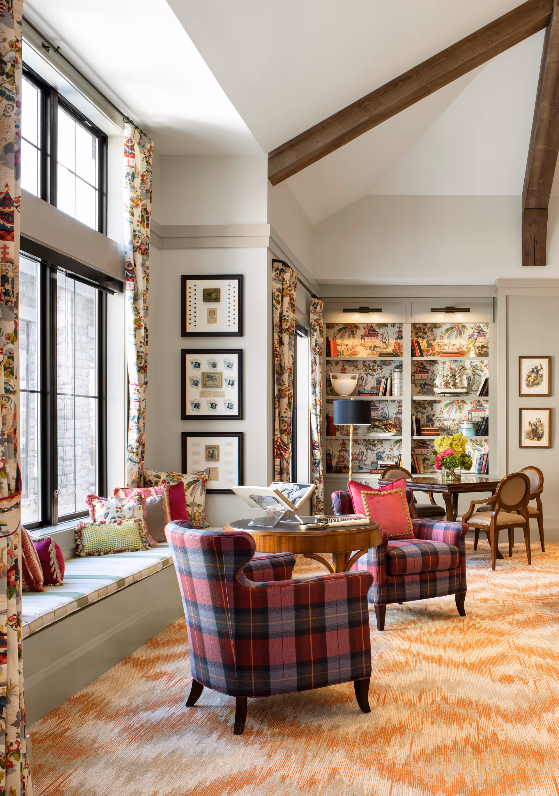 A cozy and elegant living room area with large windows featuring floral curtains, a window seat with colorful cushions, two plaid armchairs with red and purple tones around a round wooden table, and a bookshelf with decorative items and books. The room has a high ceiling with exposed wooden beams and a patterned carpet in warm tones.
