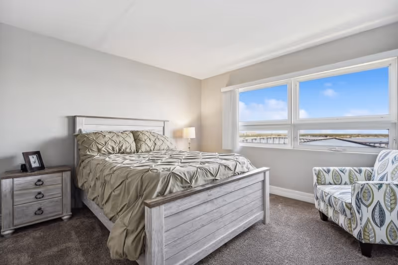 A bright bedroom with a large window showing a blue sky outside. The room features a wooden bed with a beige comforter and matching pillows, a wooden nightstand with a framed photo and a lamp, and a patterned armchair with leaf designs near the window. The walls are painted light gray and the floor is carpeted.
