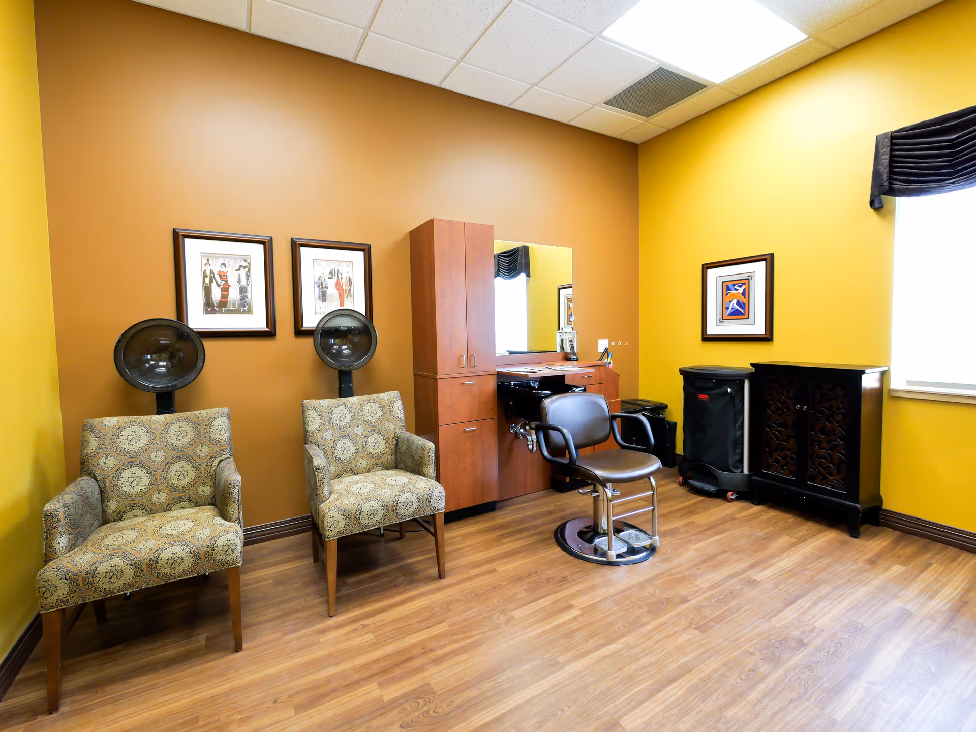 Salon-style room with two hooded hair dryers, a styling chair, cabinets, and framed artwork.