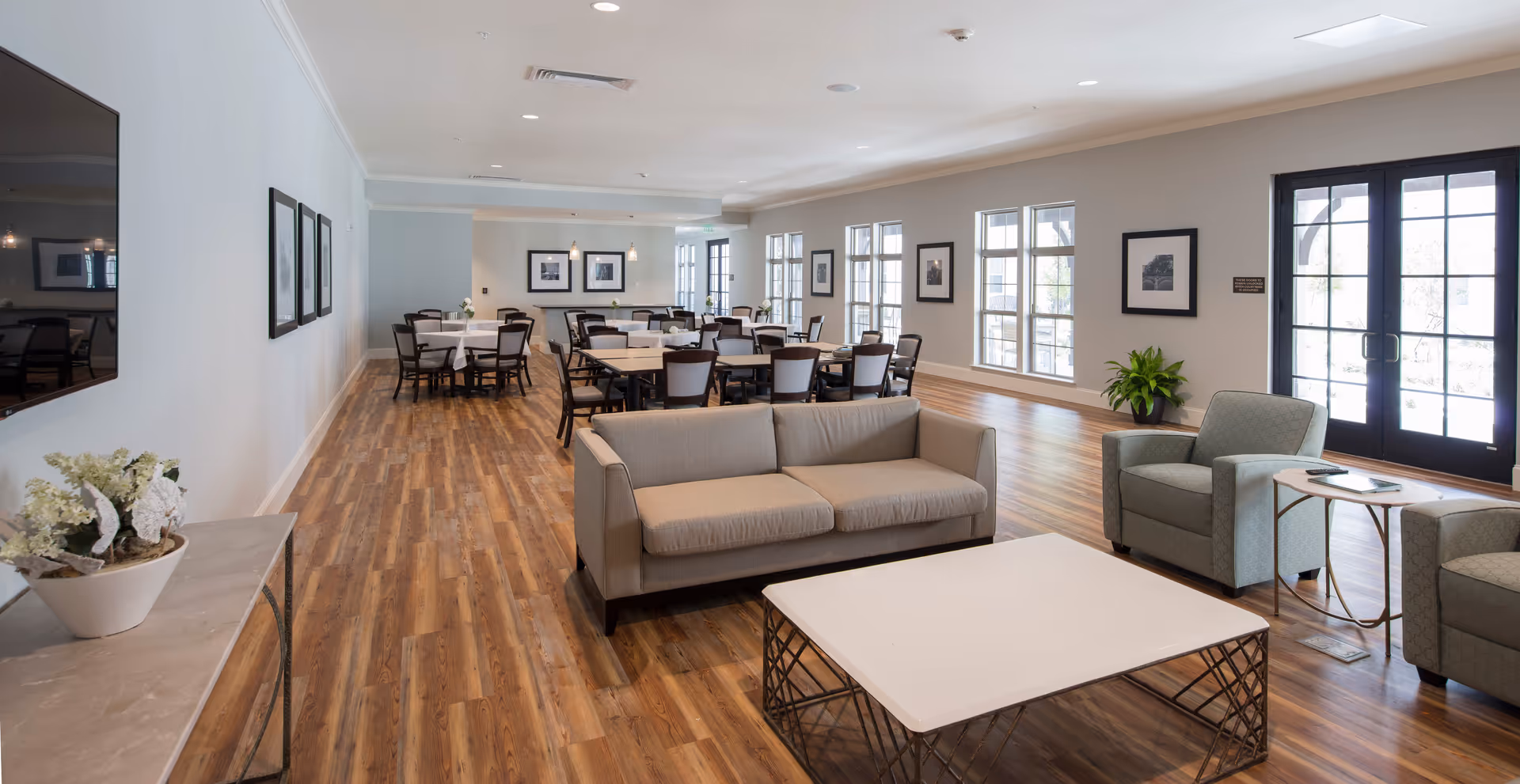 A spacious, well-lit senior living common room with sofas, armchairs, a coffee table and dining tables along a long wooden floor.