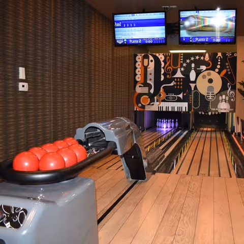 Indoor bowling alley with two lanes, red bowling balls on a ball return, and two screens displaying player scores above the lanes. The back wall features a colorful mural with musical instruments and notes.