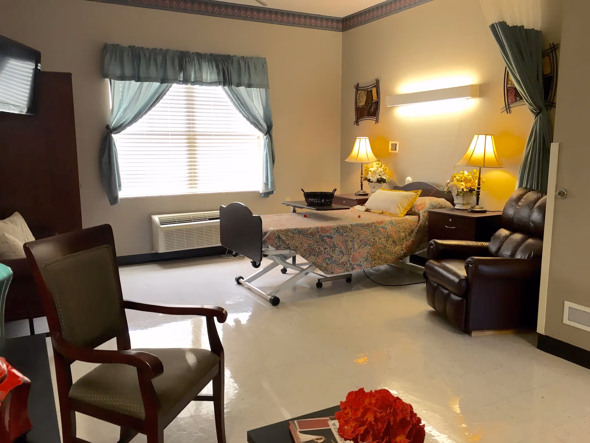 A nursing facility room with a hospital bed covered in a patterned bedspread, two bedside tables with lamps and flower arrangements, a large window with blue curtains, a brown leather recliner, a wooden chair with green upholstery, and a wall-mounted television.