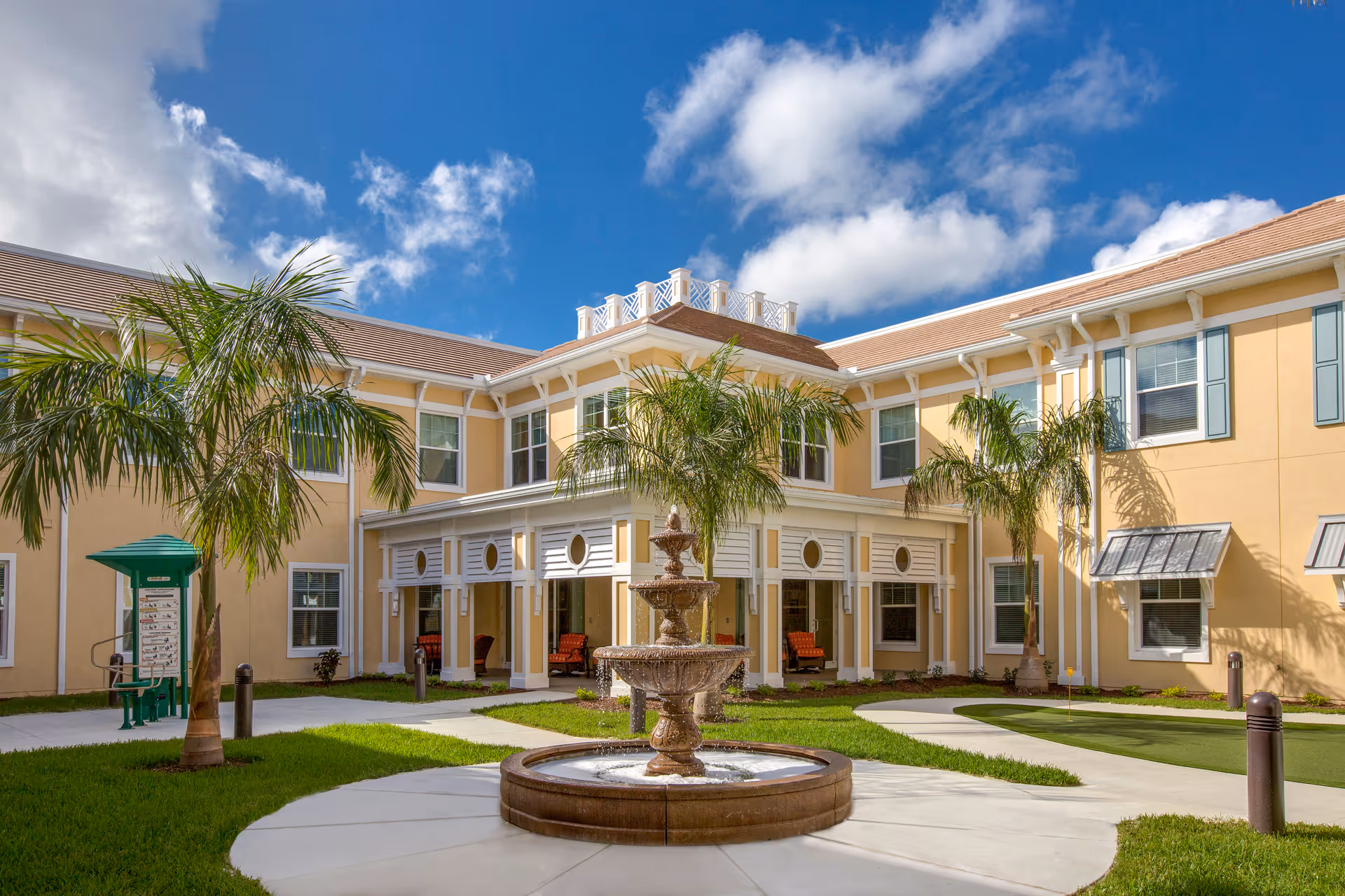 Exterior courtyard of a senior living facility with a central three-tiered water fountain, palm trees, green grass, and a two-story yellow building with white trim and multiple windows under a partly cloudy blue sky.