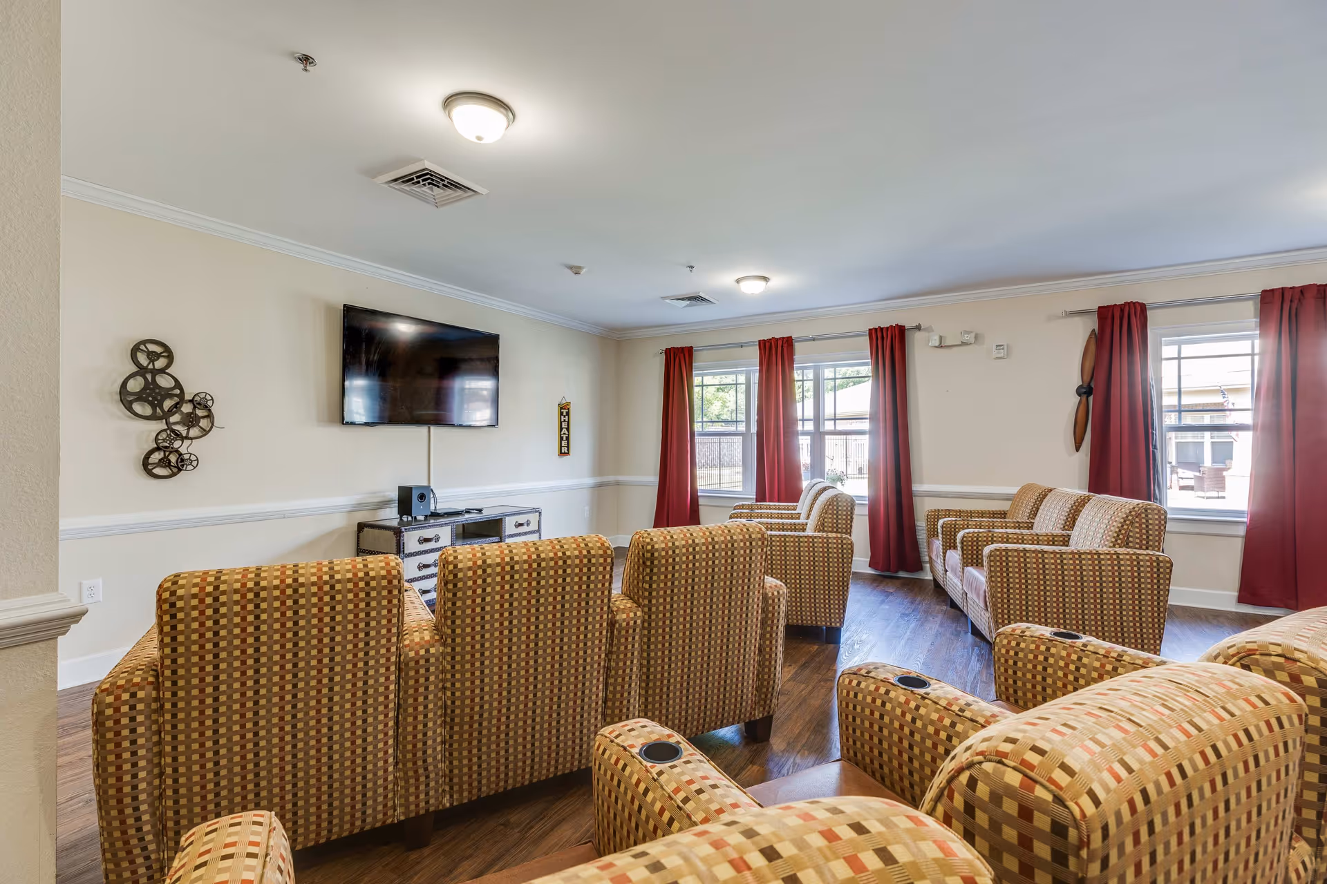 A cozy assisted living facility theater room with multiple patterned armchairs arranged in rows facing a wall-mounted flat screen TV. The room has light beige walls, red curtains on the windows, and decorative wall art including a metal film reel sculpture and a wooden propeller. The floor is wood, and the room is well-lit with ceiling lights.