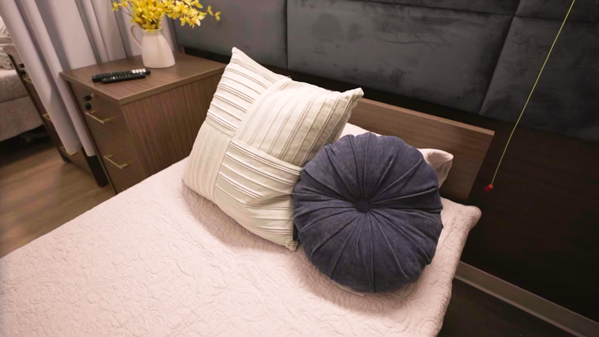 A neatly made bed with a light pink quilt and two decorative pillows, one rectangular and one round, in a bedroom. Next to the bed is a wooden nightstand with a remote control and a white vase holding yellow flowers. The room has a dark padded headboard and wooden flooring.