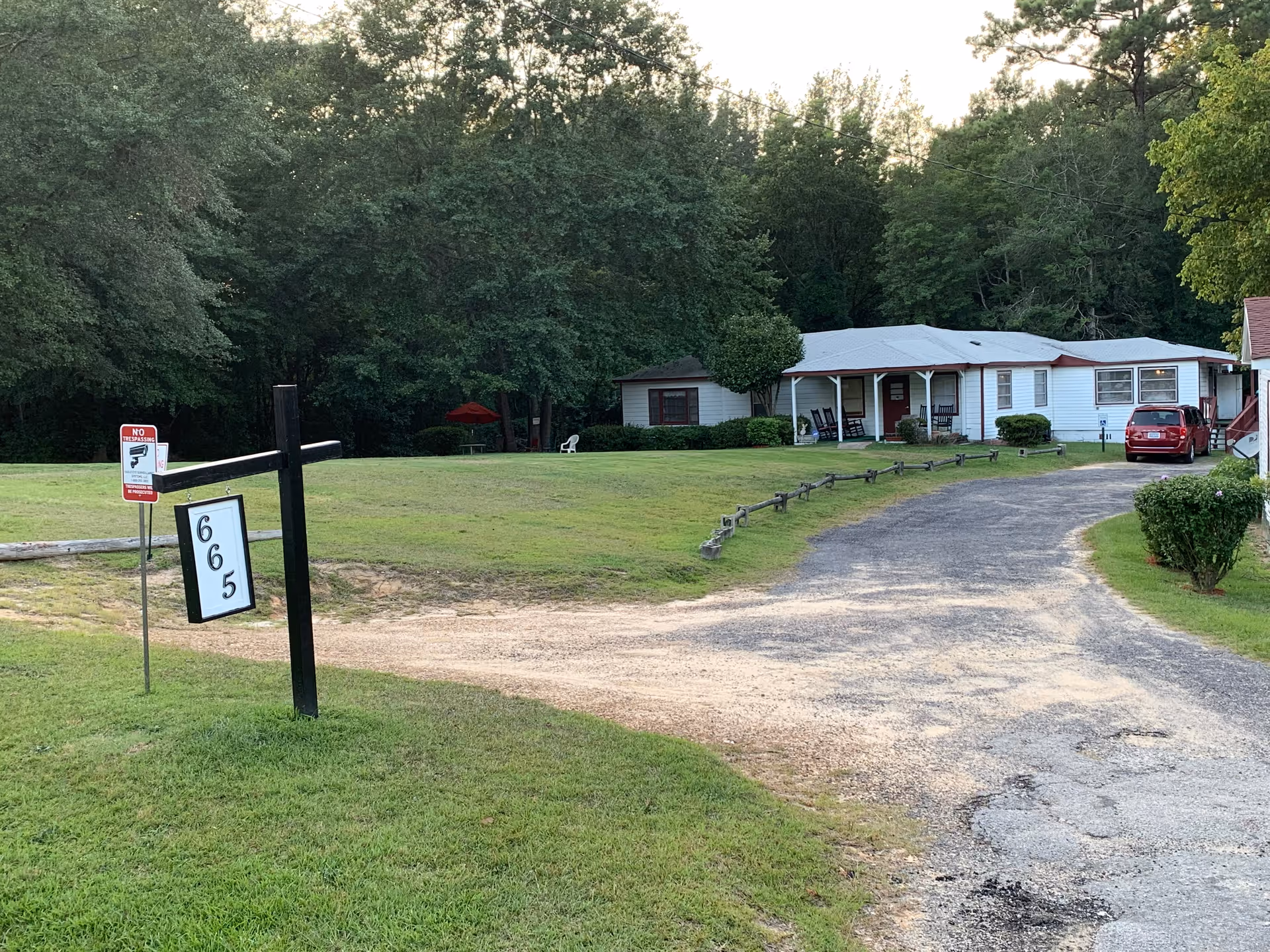 A single-story white building with a gray roof set back from a gravel driveway. The building has a covered porch with rocking chairs and is surrounded by green grass and trees. A black post near the driveway displays a sign with the number 665. There is a red car parked near the building and a 'No Trespassing' sign with a gun symbol is visible on a metal pole near the driveway.