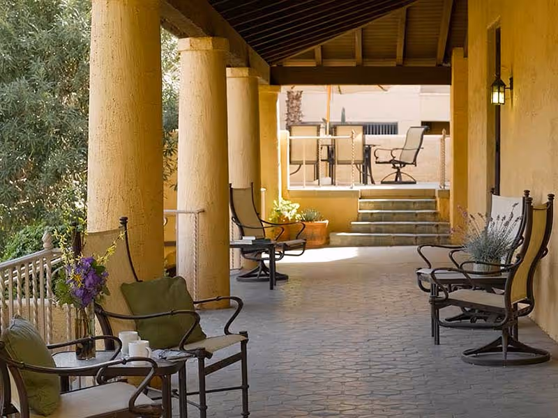 Covered outdoor patio area with cushioned chairs and small tables, potted plants, and large columns supporting the roof. Steps lead up to another seating area with a table and chairs in the background.