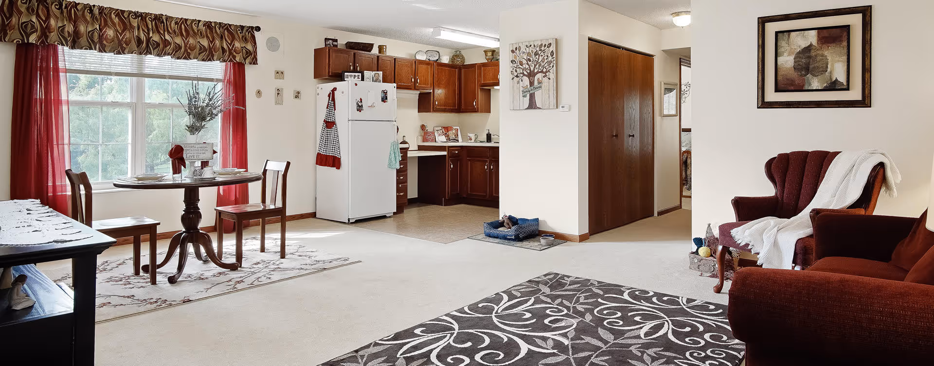 A bright and cozy living space featuring a dining area with a round wooden table and two chairs near a large window with red curtains. The kitchen area has wooden cabinets, a white refrigerator, and various kitchen items. The living room area includes a red armchair and sofa with a white throw blanket, a patterned rug, and framed artwork on the walls. There is also a pet bed on the floor near a hallway with wooden doors.