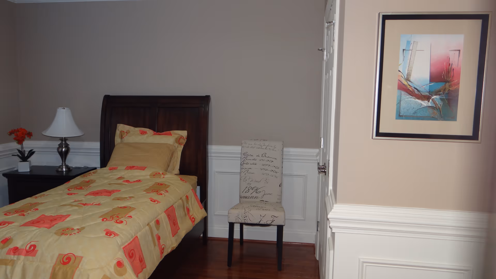 A single-bed bedroom with a yellow-and-pink patterned bedspread, wooden headboard and nightstand with a lamp and flowers, plus an upholstered chair against a paneled wall.