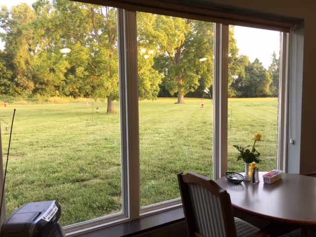 View from a dining area through large floor-to-ceiling windows onto a grassy lawn with trees.