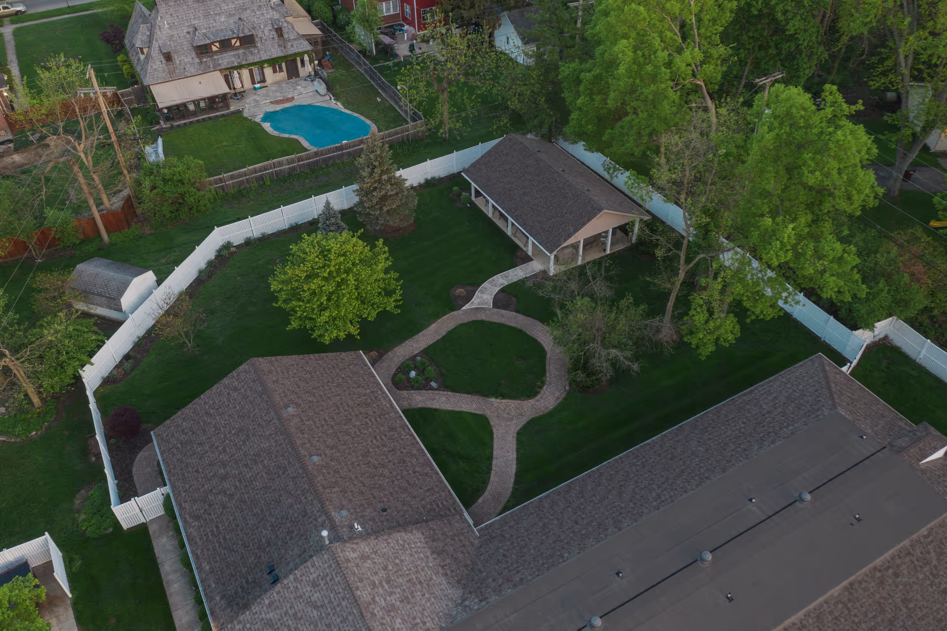 Aerial view of a fenced courtyard with winding brick paths, a small pavilion, trees, and surrounding building roofs.
