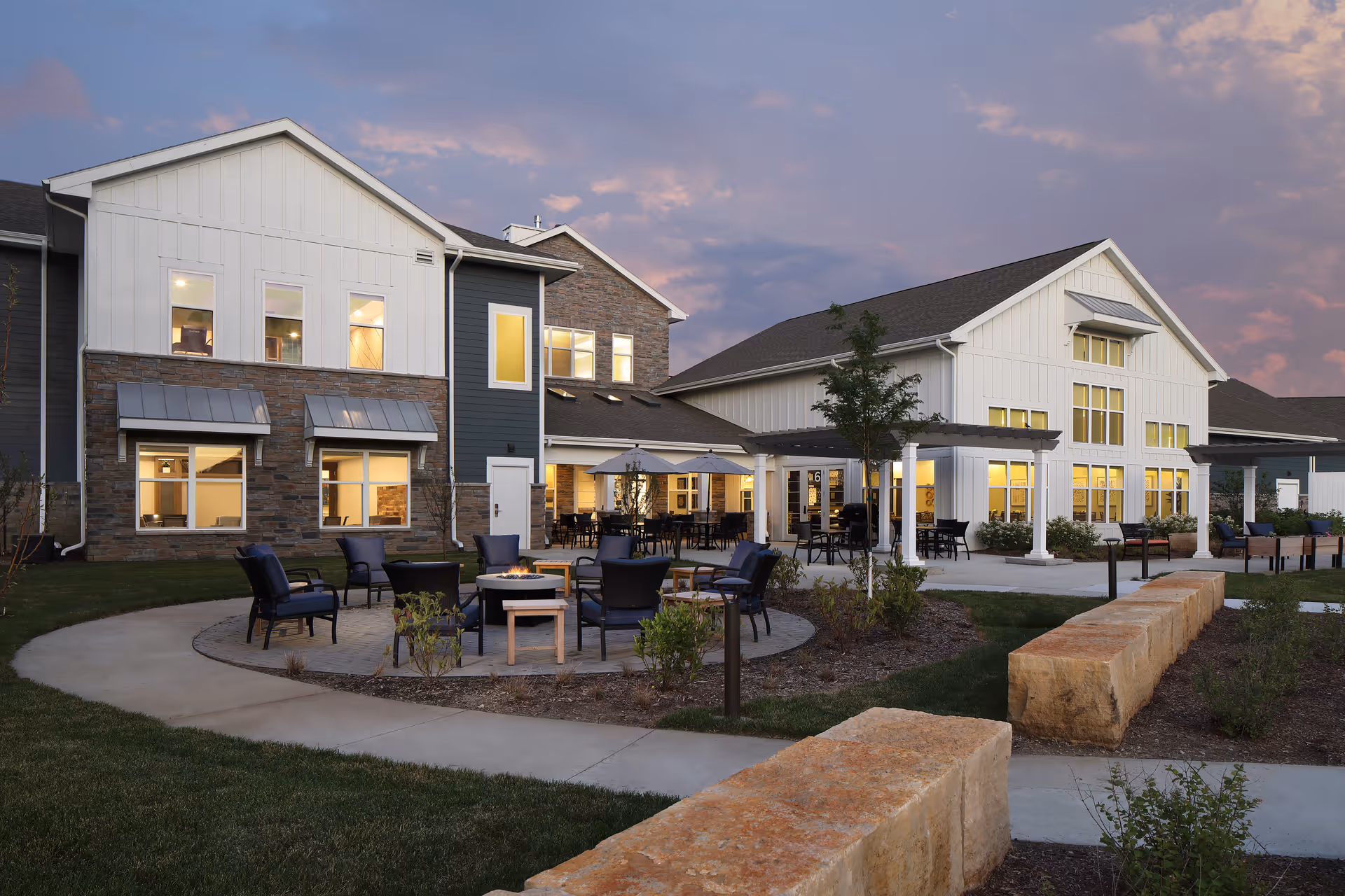 Outdoor seating area at CedarStone Senior Living with chairs arranged around a fire pit, surrounded by a paved walkway and landscaped garden beds, adjacent to a large two-story building with lit windows during dusk.