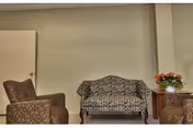 A small seating area with a patterned loveseat and two matching armchairs arranged around a wooden side table with a vase of flowers, set against a plain beige wall with a closed door on the left.