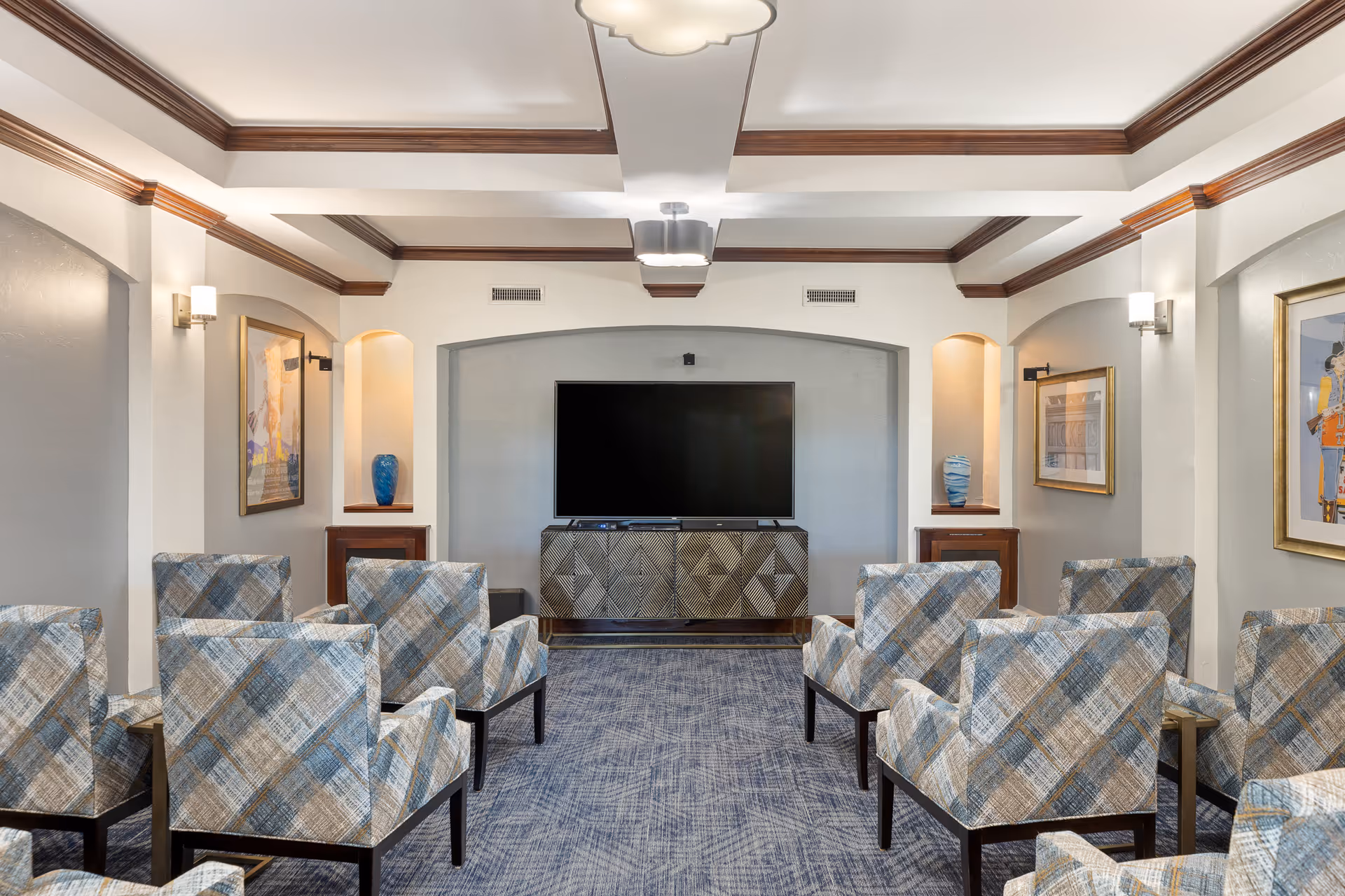 A cozy living room or common area with multiple patterned armchairs arranged in rows facing a large flat-screen TV mounted on a decorative cabinet. The room features a blue carpet, white walls with wooden crown molding, built-in wall niches with blue vases, and framed artwork on the walls. Ceiling lights provide soft illumination.