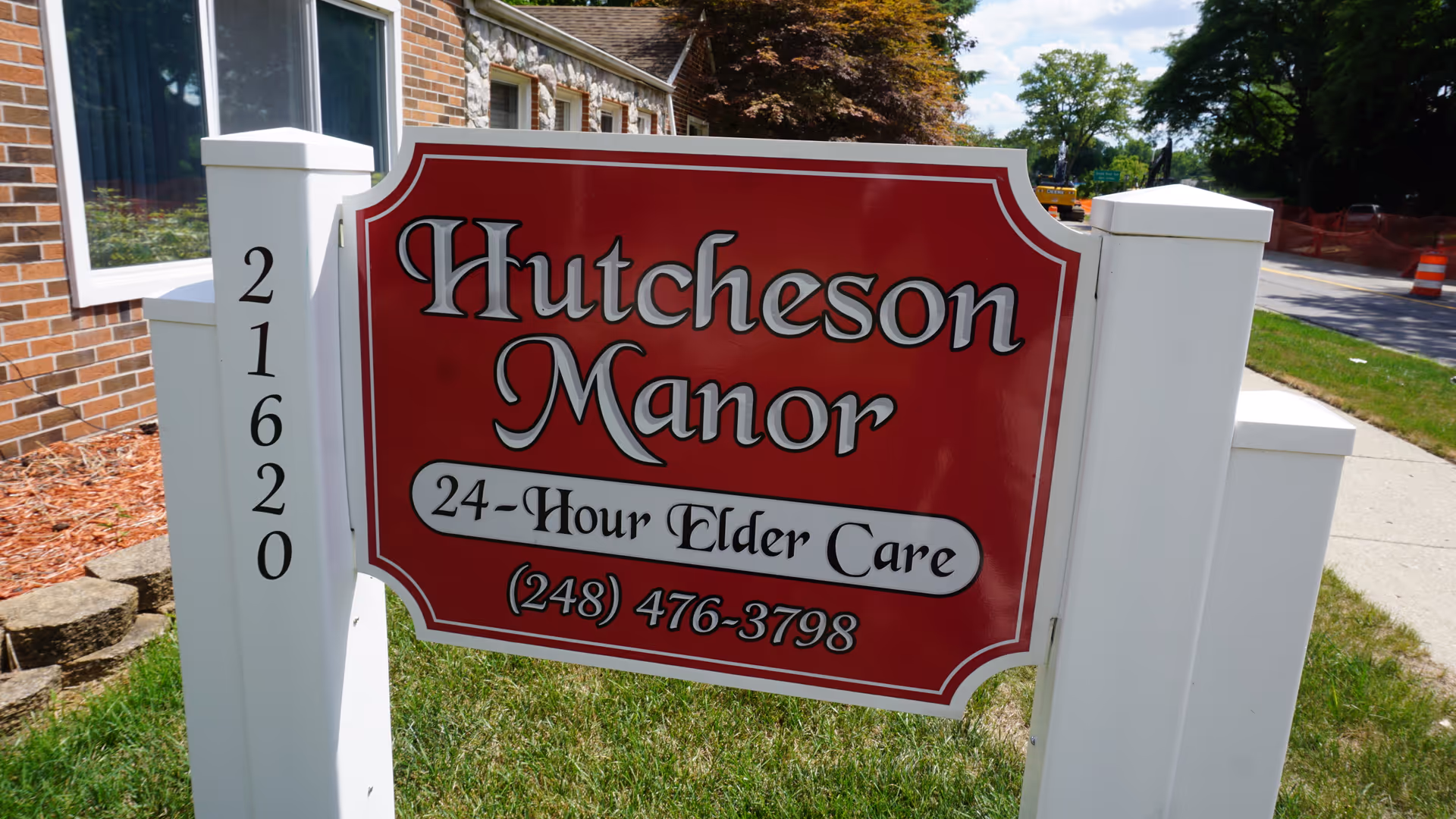 A red sign reading 'Hutcheson Manor 24-Hour Elder Care (248) 476-3798' mounted on white posts in front of a brick building and lawn.