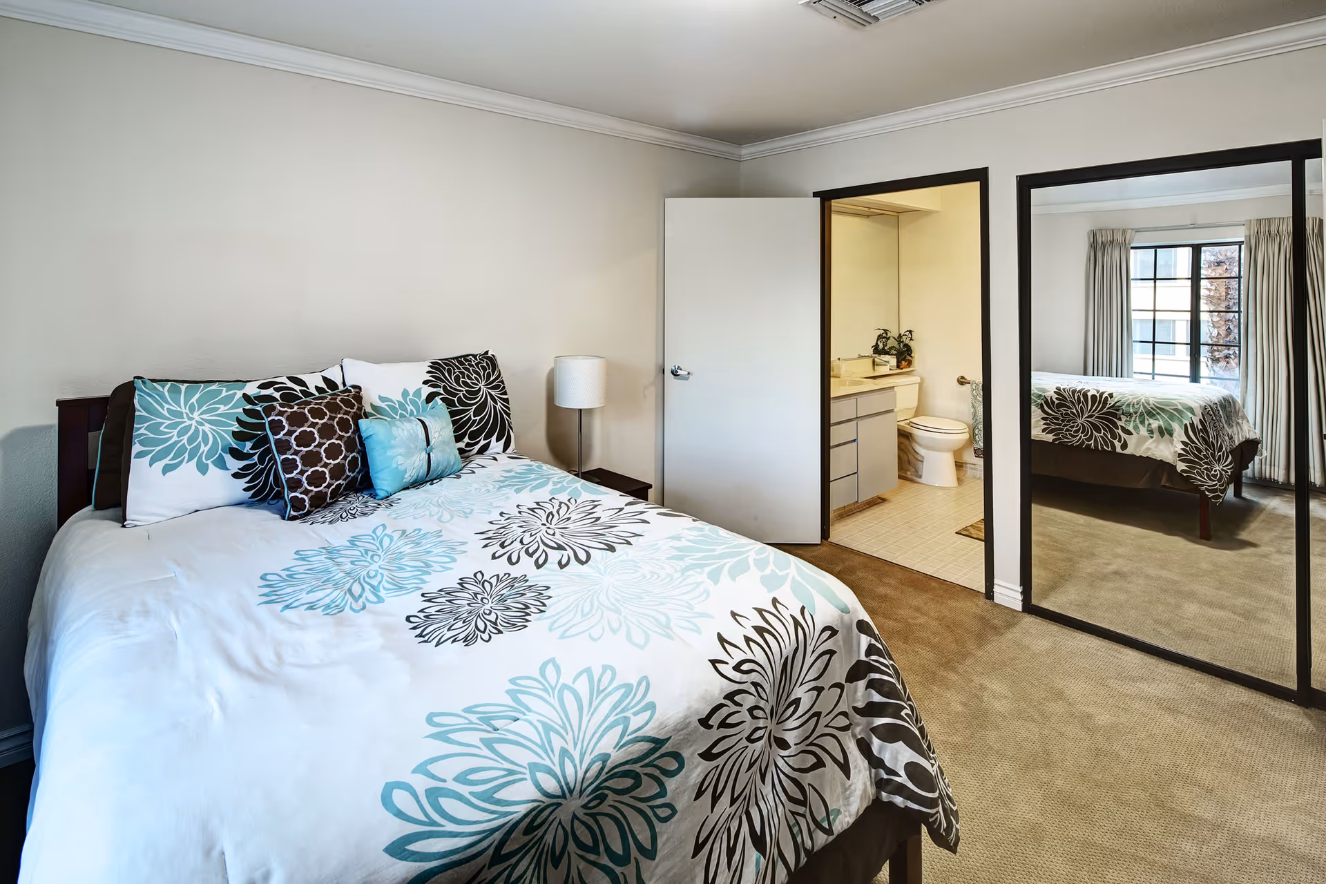 A bedroom with a bed covered in a white, teal, and brown floral patterned comforter and multiple pillows. There is a small bedside table with a lamp next to the bed. An open door reveals a bathroom with a toilet, sink, and mirror. A large mirrored closet door reflects the bed and a window with curtains.