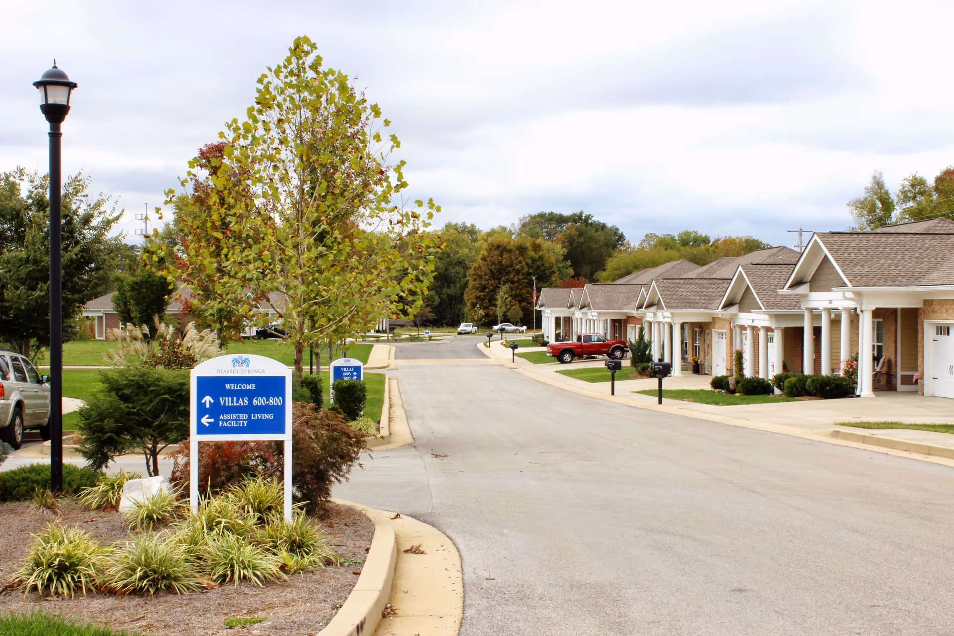 A quiet street in a senior living community with single-story villas lined up on the right side. There are small trees and landscaped bushes along the street, a lamp post on the left, and a blue sign indicating directions to villas and an assisted living facility. Cars are parked near the villas and the sky is overcast.