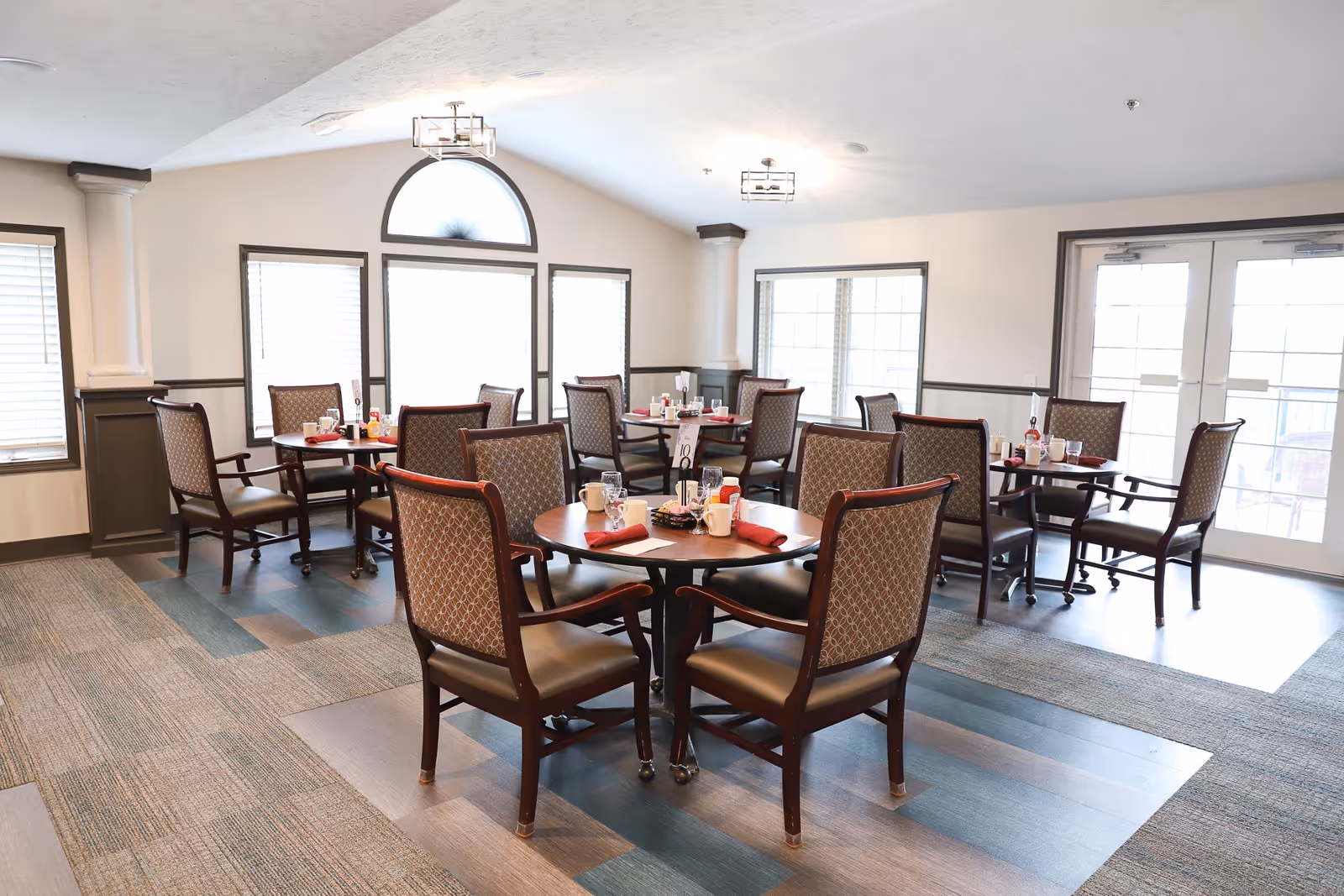 A bright dining room with several round tables, each surrounded by four upholstered chairs. The tables are set with cups, glasses, napkins, and condiments. Large windows and glass doors allow natural light to fill the room, which has a high ceiling and a patterned carpeted floor.