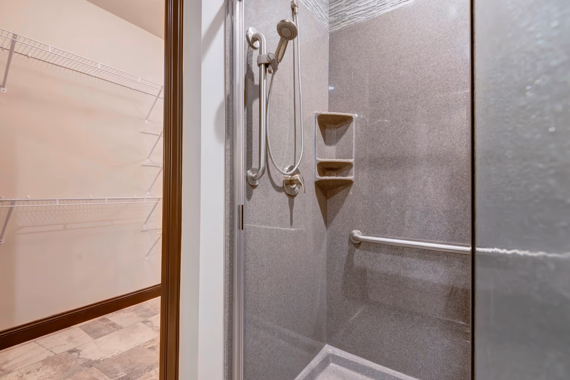 Interior view of a shower area with a handheld showerhead, grab bars, and built-in shelves. Adjacent to the shower is a walk-in closet with wire shelving and a tiled floor.