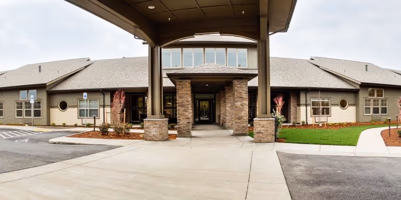 Front exterior view of Waterhouse Ridge Memory Care facility showing a covered entrance with stone pillars, a driveway, parking spaces including handicapped spots, and landscaped areas with grass and small trees.
