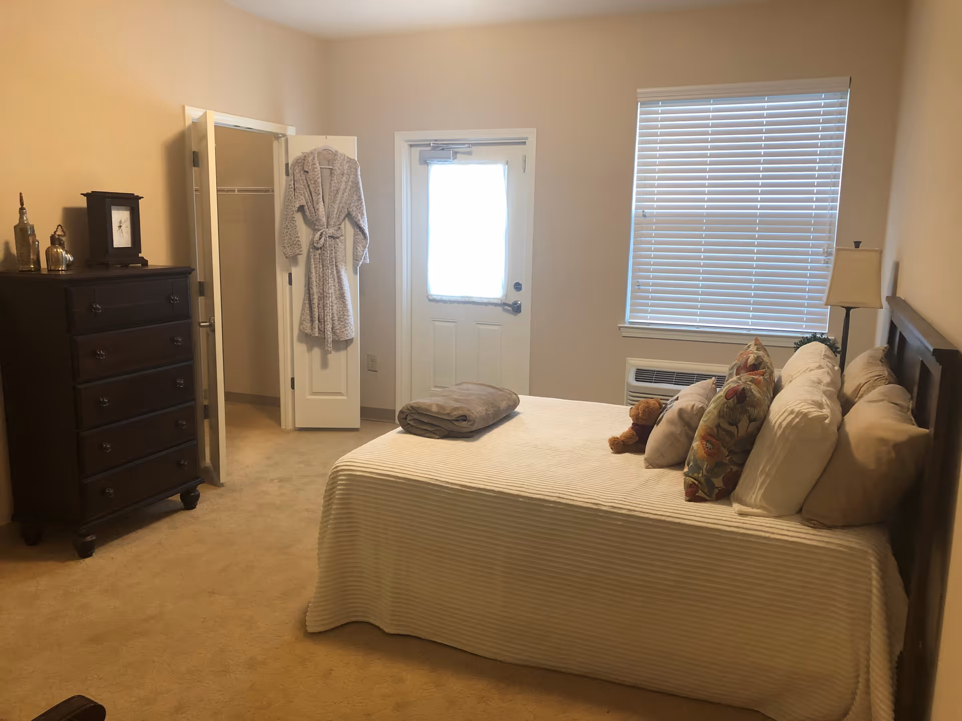 A bedroom with a bed covered in a white bedspread and multiple pillows, including floral and plain ones. A small teddy bear is placed on the bed. To the left, there is a dark wooden dresser with decorative items on top and an open closet door with a robe hanging on it. A window with closed blinds and a door with a frosted glass panel are visible on the far wall. A floor lamp stands next to the bed.
