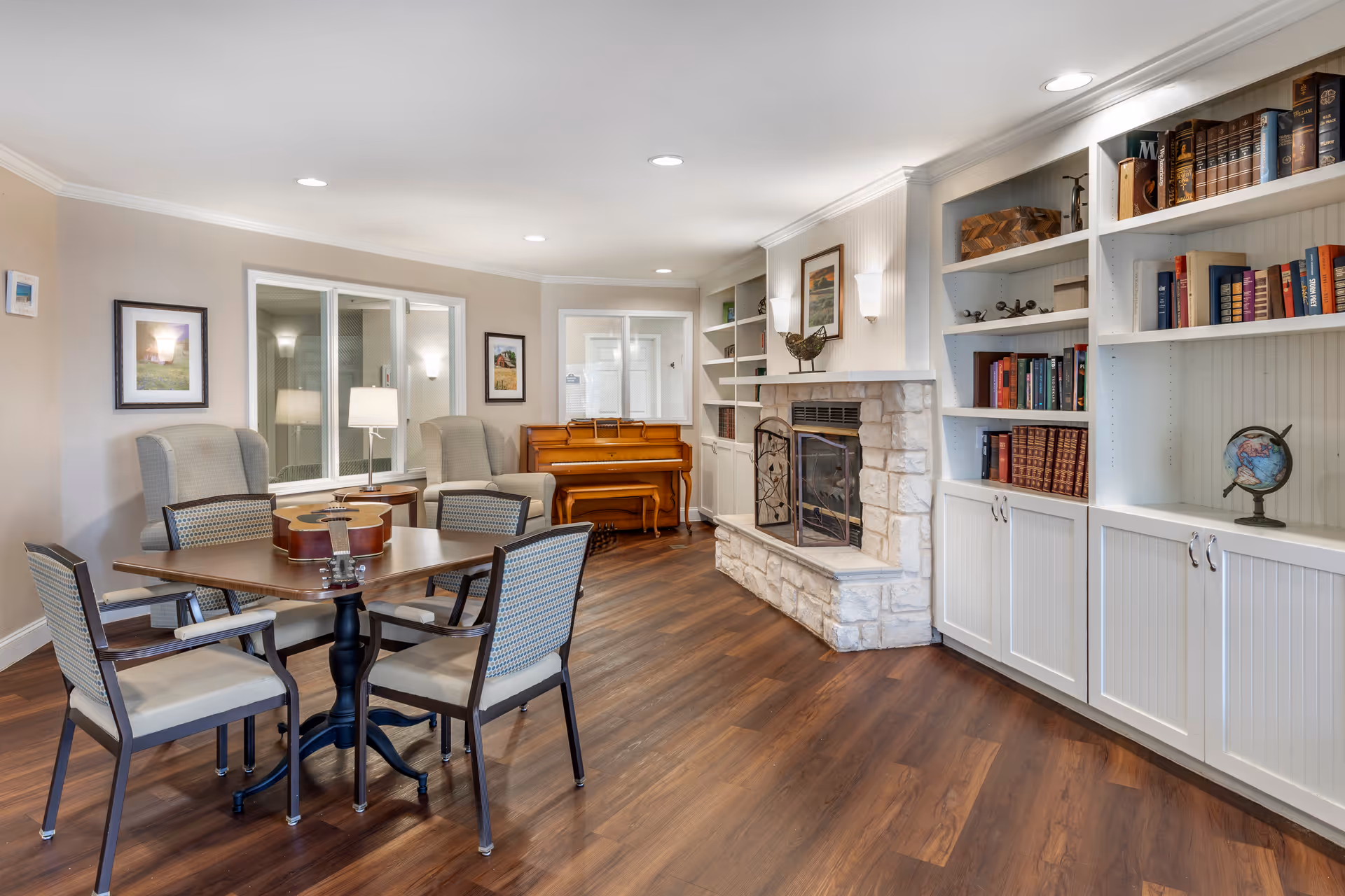 A cozy common area with a wooden floor, featuring a square table with four chairs around it and a guitar placed on the table. There are two upholstered armchairs with a small round side table and lamp between them. In the background, there is a wooden piano against the wall. To the right, a stone fireplace with a decorative screen is flanked by built-in white bookshelves filled with books and decorative items, including a globe. The walls are light-colored with framed pictures and recessed ceiling lights providing illumination.